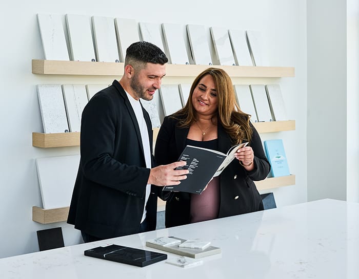 Two Stone Design Concepts team members reviewing a design catalog together in a modern showroom, with samples displayed on wall shelves.