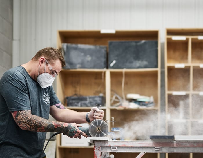 Skilled Stone Design Concepts craftsman using precision tools to shape stone, wearing a mask for safety, in a workshop with shelves in the background.